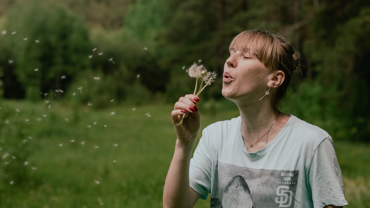 A woman in a t-shirt blows dandelion seeds in a lush green field during summer.